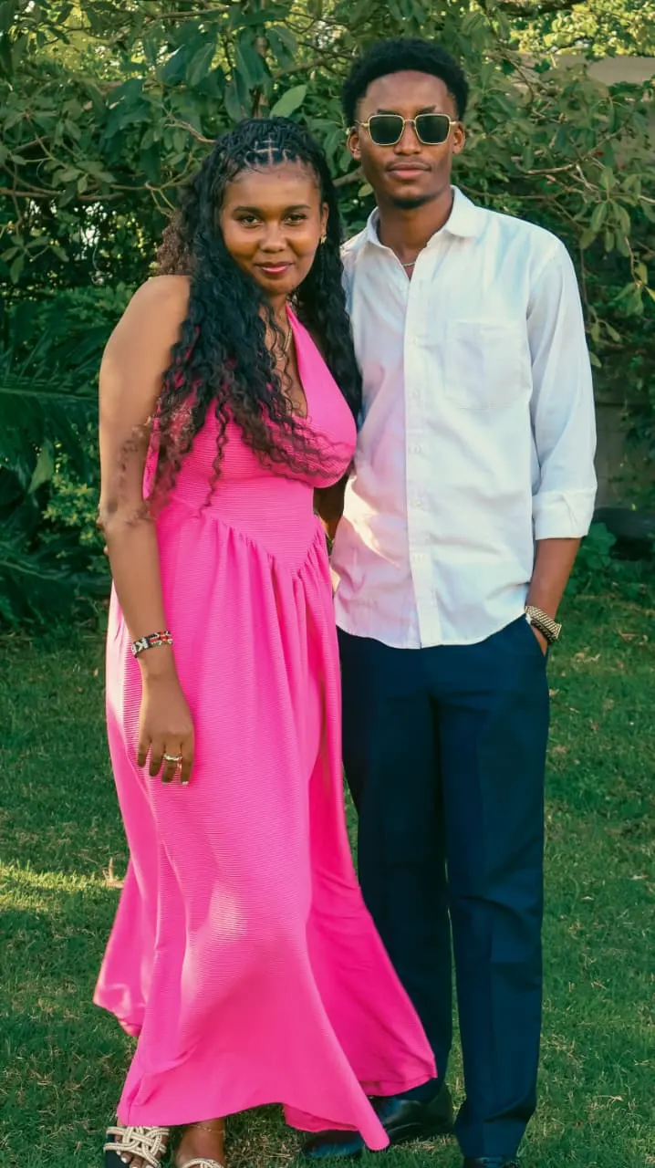 A couple dressed for celebration standing close together in a sunlit garden, she in pink, he in white