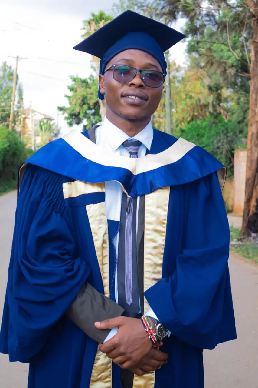 A graduate in a navy gown with gold trim, cap on, holding his certificate on a tree-lined road