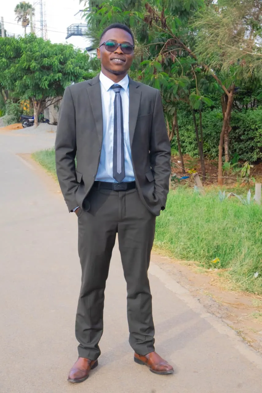 The graduate in a pressed brown suit standing tall on a quiet tree-lined road before the ceremony